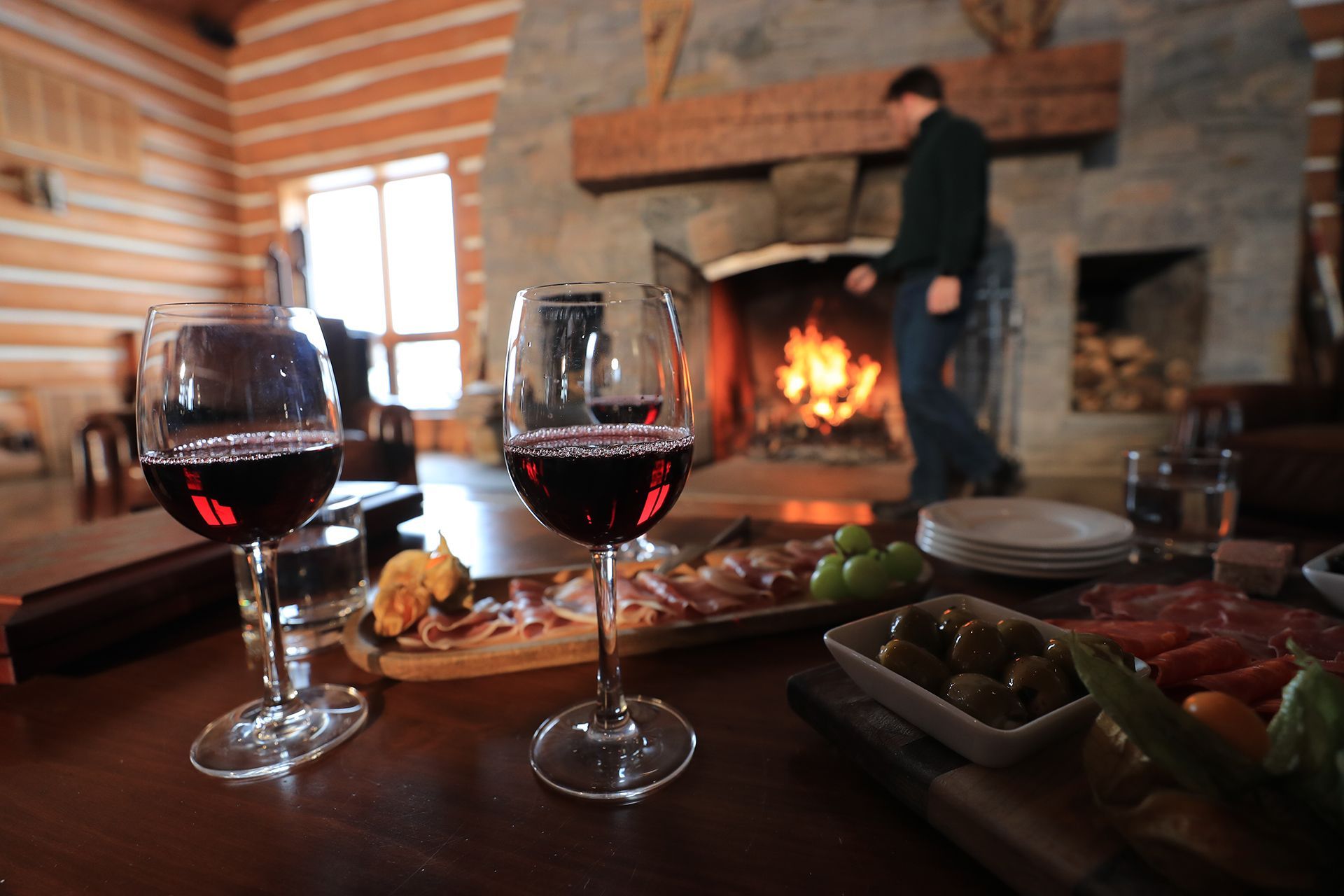 glasses of red wine and plates of charcuterie and olives sit on a wood table in front of a fireplace