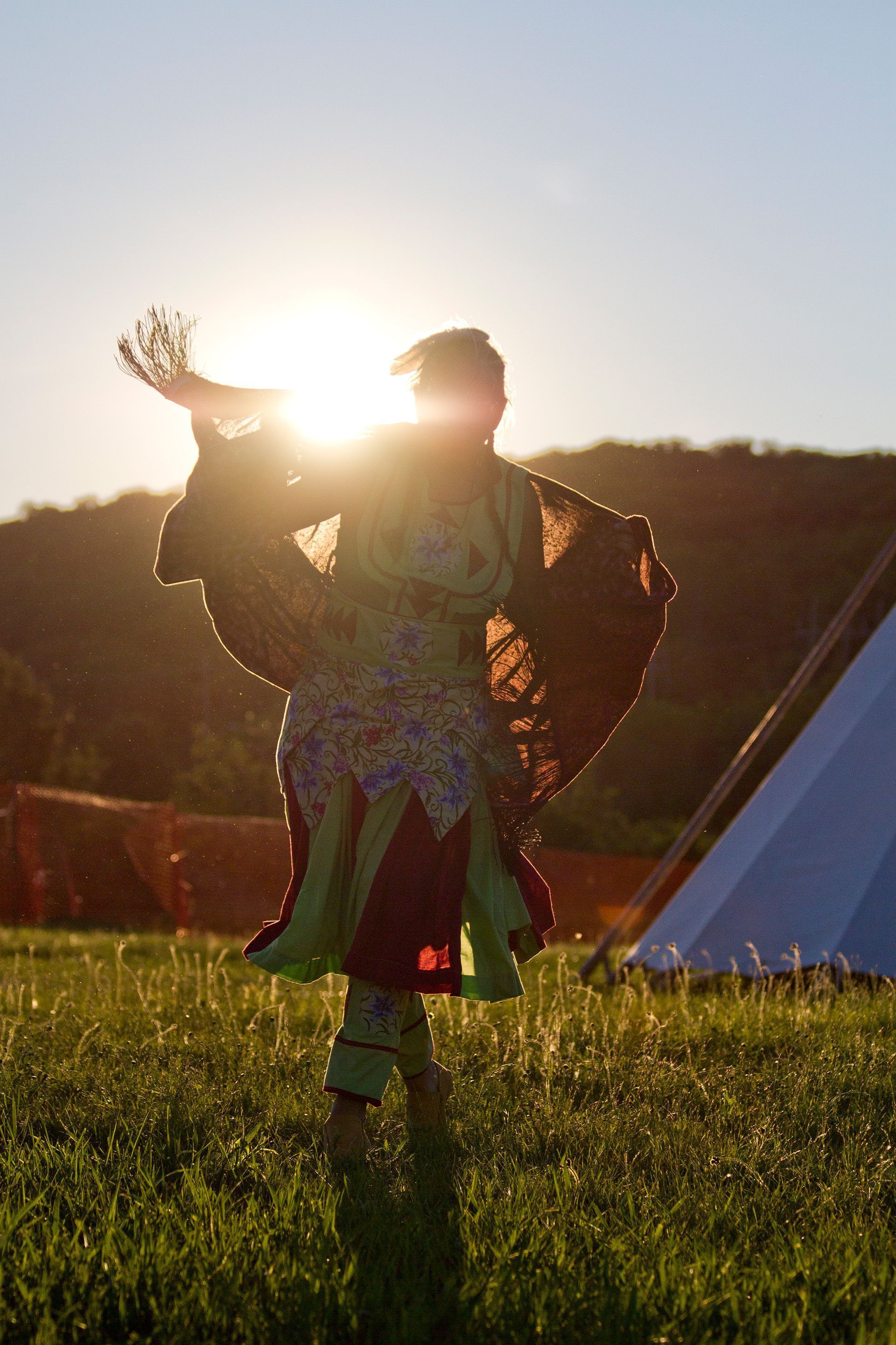 A person wearing regalia dances while the sun shines behind her.