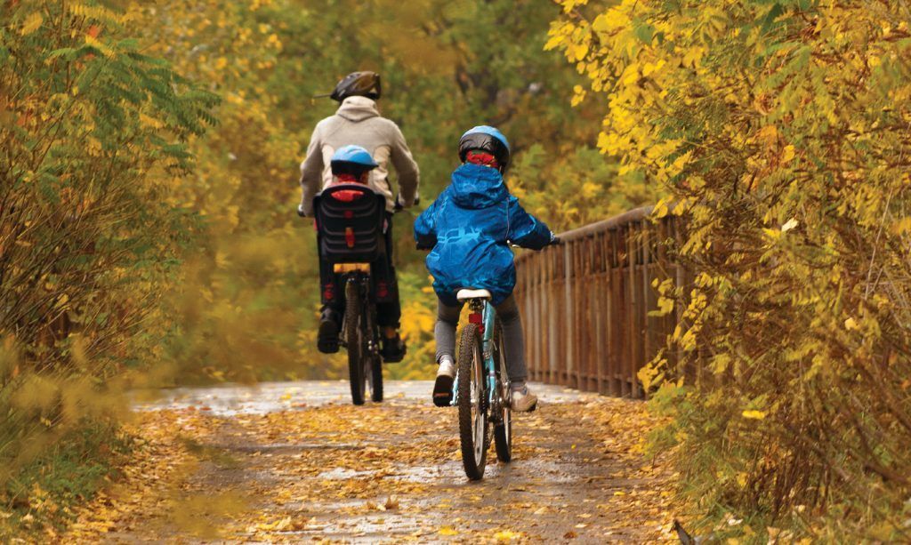 A mother and her two kids bike across a bridge surrounded by fall colours.