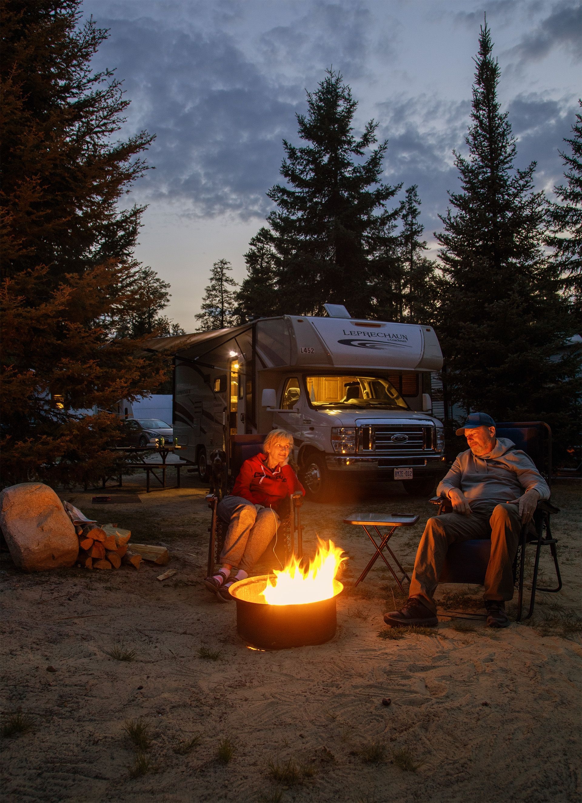 Two people sit in front of a campfire by their RV