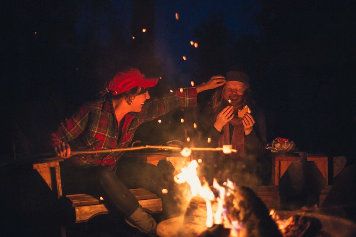 Two women sitting at a campfire, laughing as they roast marshmallows and make s'mores together