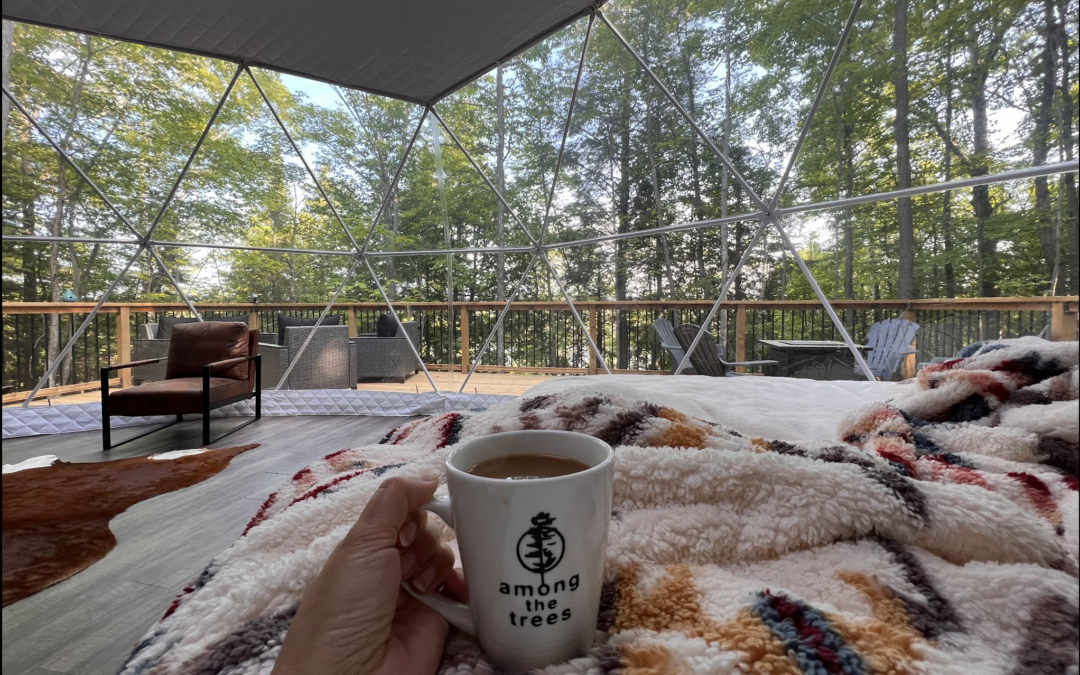 Guest at Among the Trees Glamping Domes lies curled up in bed with a steaming cup of coffee, looking out the panoramic windows at a view of Boreal forest on Birch Island, Ontario